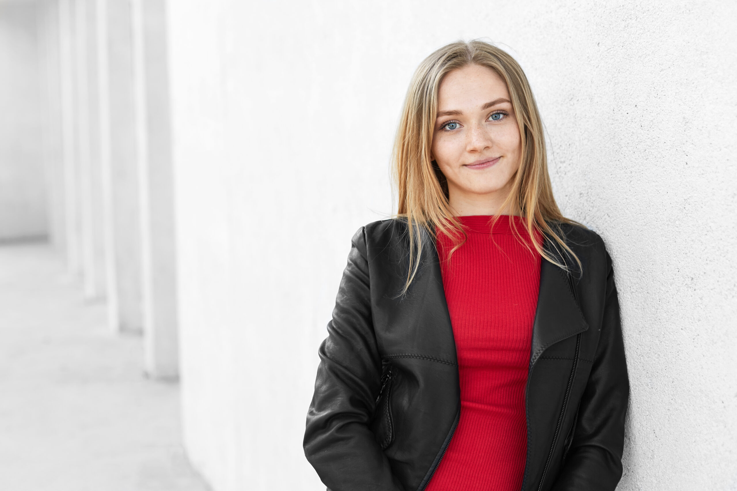 Horizontal portrait of beautiful fashionable teenage girl with fair hair, freckled face dressed in trendy clothes standing near white wall having pensive expression. Woman with charming appearance