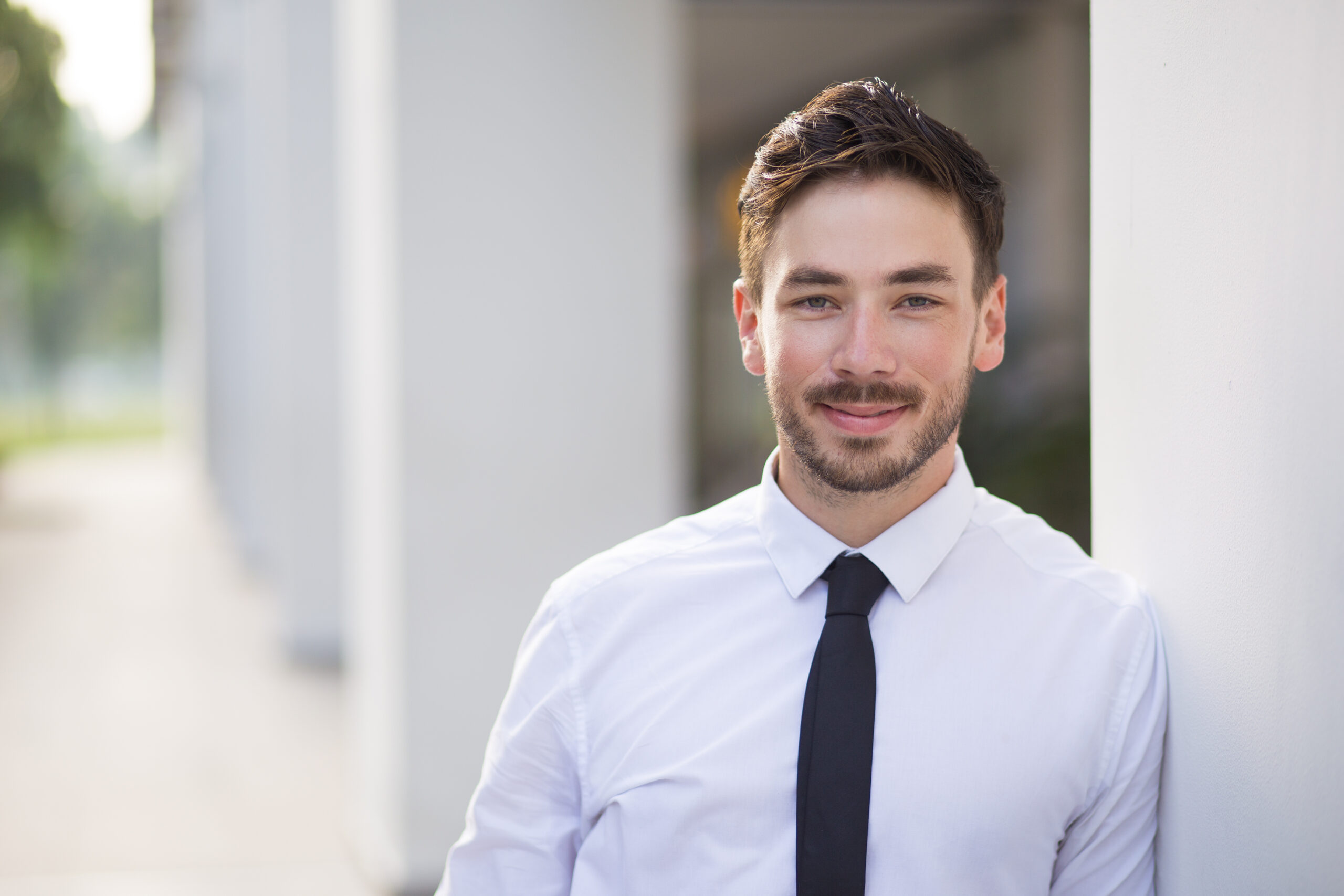 Smiling young entrepreneur posing for camera and standing in street. Positive businessman leaning on column and looking at camera. Business portrait concept